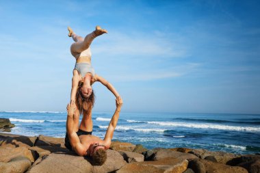 Genç ve zinde bir çift deniz sahilindeki spa merkezinde akro yoga yapıyor. Eş ayaklı aktif bir kadın, amuda kalkarak akroyoga pozunda dengede duruyor. Sağlıklı bir yaşam tarzı. Aile tatilinde insanlar açık havada spor yapıyor..