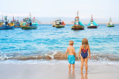 Happy family - kids walk along sea surf on sand beach, look at traditional fisher boats. Pantai Tanjung Papuma, Jamber, Java, Indonesia. Popular travel destination. Summer vacation with kids. clipart