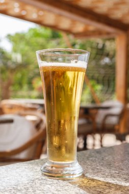 Yazlık bir kafede masanın üzerinde hafif bira olan bir bardağın dikey resmi. Sıcak bir günde hafif alkollü içecekler. Tatil köyündeki bar.