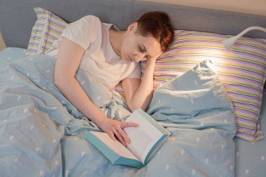 Young red-haired woman fell asleep in bed while reading before bedtime. Woman fell asleep over an open book in the light of the bedside lamp.