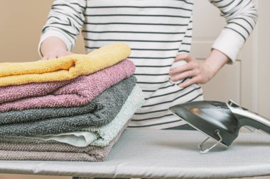 Woman stands by an ironing board with an iron and is holding a stack of ironed, clean terry towels. Housework, ironing of the washed linen. The housekeeper is engaged in household chores.