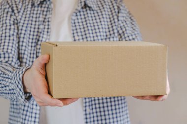 Courier in a check shirt holds out a sealed cardboard box. Delivery of online orders. The delivery clerk brought the packaged order. Space for text.