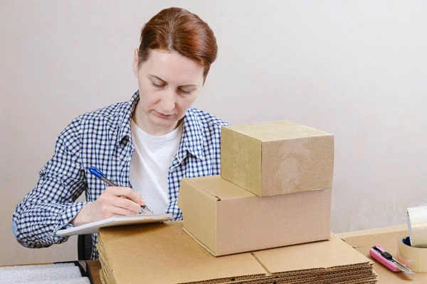 Business woman is packing orders of her online store. A woman makes notes in a notebook, keeps track of orders. Small business, work from home, order processing and packing.