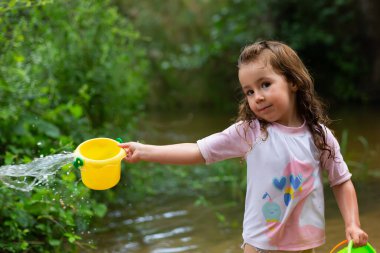 Küçük kız nehirde plastik kovalarla oynuyor. Mayolu ve tişörtlü kız. Yazın çocuk fotoğrafı..