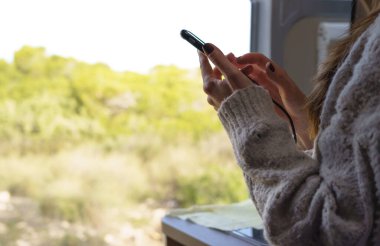 Bir karavanın içinde akıllı telefonunu kullanan bir kadının yatay görüntüsü. Güzel bir günün tadını çıkarmak için kapı açık..