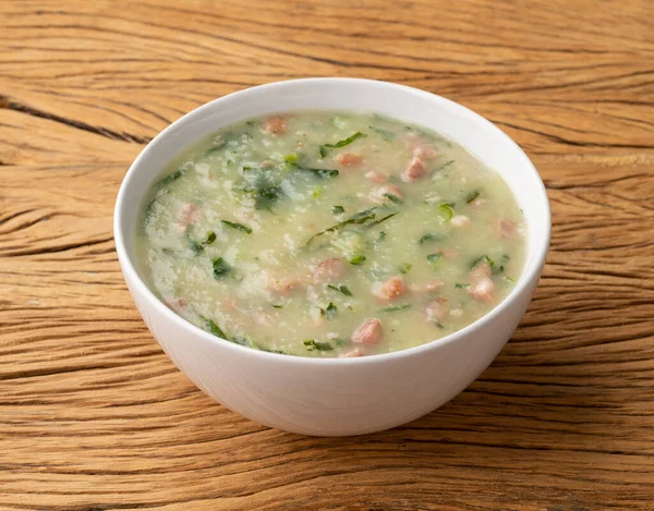 Traditional portuguese green soup with potato, green cabbage and sausage in a bowl over wooden table.
