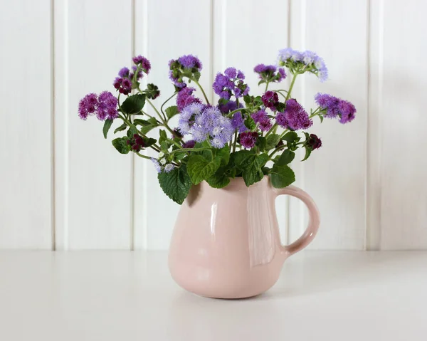 Pink jug with a purple bouquet of ageratum. — Stock Photo, Image