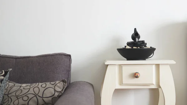 Front view of a decorative zen water fountain on top of a small white table next to a gray couch. A white wall as a background