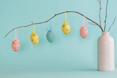 Süslü Paskalya yumurtaları beyaz seramik vazoda asılı ve mavi arka planda iki mavi yumurta var. 