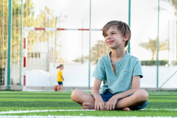 Calm boy sitting on green lawn with ball and looking seriously away