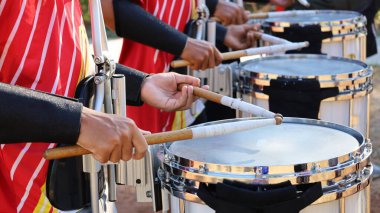 Çubuklu ve davullu eller. Kırmızı gömlekli adamlar açık hava orkestrasında trampet çalıyorlar. Seçici odak