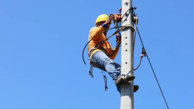 Bir elektrikçi beton bir direğe kablo yerleştirir. İşçiler kemerleri güvenlik direklerine asar ve düşük voltajlı kabloları fotokopi alanı olan açık bir gökyüzüne beton direklere yerleştirirler. Seçici odak