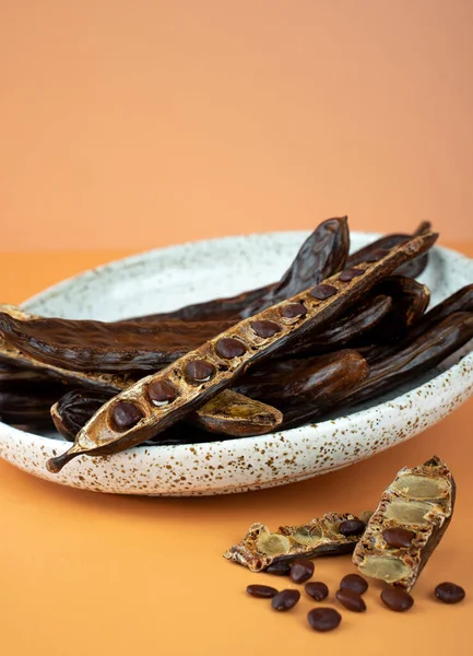 Organic carob pods, seeds in a ceramic plate on a beige background, locust bean healthy food, Ceratonia siliqua harnup. Natural vegan eating. creative food background. Copy space. Top view