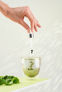 Kadın eli, bir bardak suya bir damlatıcıyla sıvı klorofil döker. Yeşil masada sıvı klorofil ve taze bitkiler var. Yeşil arka plan, doğal güneş ışığı, dikey. Süper yemek ve sağlıklı.