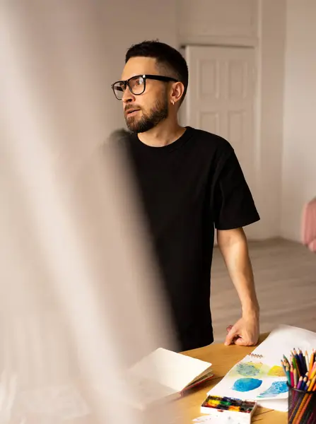 The artist is trying to figure out how to create a masterpiece. A young man standing over a table in the workshop. Process of finding inspiration, working in the artist\'s studio, blurred foreground.