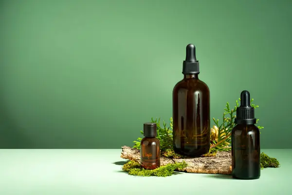 A set of brown glass bottles, moss and tree bark on a green background. Oil with serum for skin and hair care. Glass bottle of essential body oil with a dropper. Self-care and wellness. Copy space.