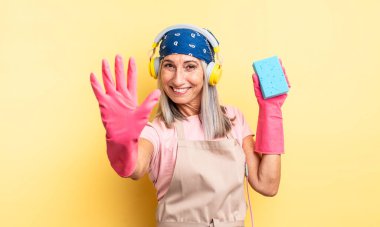 Orta yaşlı güzel bir kadın gülümsüyor ve arkadaş canlısı görünüyor, beş numarayı gösteriyor. Temizleyici.