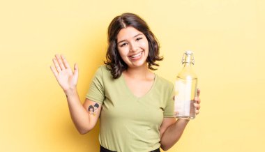 Genç İspanyol kadın mutlu bir şekilde gülümsüyor, el sallıyor, sizi selamlıyor ve selamlıyor. su şişesi kavramı