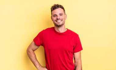 Genç, yakışıklı İspanyol adam mutlu bir şekilde gülümsüyor, eli kalçasında ve kendinden emin, olumlu, gururlu ve arkadaş canlısı.