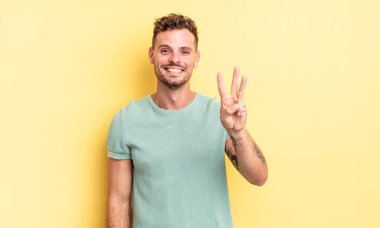 Genç, yakışıklı İspanyol adam gülümsüyor ve arkadaş canlısı görünüyor, üç ya da üç numarayı gösteriyor, eli önde, geriye doğru sayıyor.