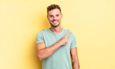 Genç, yakışıklı İspanyol adam neşeyle gülümsüyor, mutlu hissediyor ve kenarı ve yukarıyı işaret ediyor, fotokopi uzayında nesne gösteriyor.