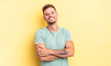 Genç, yakışıklı İspanyol adam kollarını kavuşturup mutlu bir şekilde gülüyor, rahat, pozitif ve tatmin edici bir pozla.