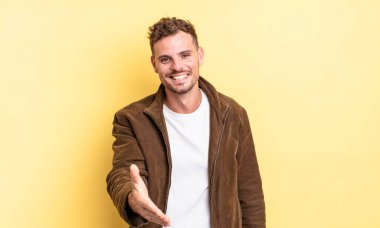 Genç, yakışıklı İspanyol adam gülümsüyor, mutlu görünüyor, kendinden emin ve arkadaş canlısı, bir anlaşmaya varmak için el sıkışmayı teklif ediyor, işbirliği yapıyor.