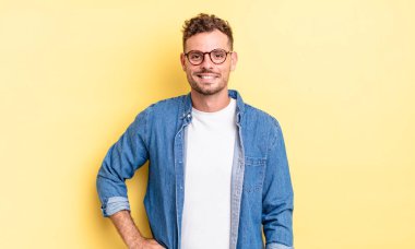 Genç, yakışıklı İspanyol adam mutlu bir şekilde gülümsüyor, eli kalçasında ve kendinden emin, olumlu, gururlu ve arkadaş canlısı.