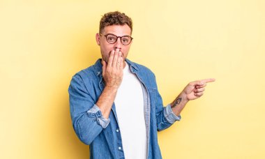 Genç, yakışıklı İspanyol adam kendini mutlu, şok olmuş ve şaşırmış hissediyor. Ağzını eliyle kapatıyor ve yanal kopya alanını işaret ediyor.