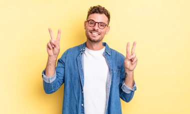 Genç, yakışıklı İspanyol adam gülümsüyor ve mutlu görünüyor, arkadaş canlısı ve memnun, iki eliyle zafer veya barış işareti yapıyor.