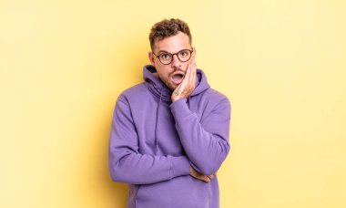 Genç, yakışıklı İspanyol bir adam şok ve inançsızlıkla ağzı açık, elleri yanağında ve kolları çaprazlanmış, sersemlemiş ve şaşırmış hissediyor.