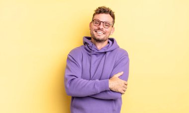 Genç, yakışıklı İspanyol adam kollarını kavuşturup mutlu bir şekilde gülüyor, rahat, pozitif ve tatmin edici bir pozla.