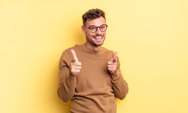 Genç, yakışıklı İspanyol adam pozitif, başarılı, mutlu bir tavırla gülümsüyor kamerayı işaret ediyor, elleriyle silah işareti yapıyor.