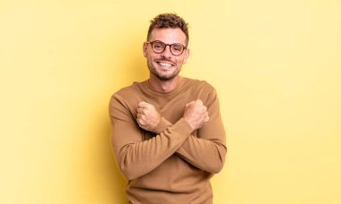 Genç, yakışıklı İspanyol adam sevinçle gülümsüyor ve kutluyor, yumruklarını sıkıp kollarını kavuşturuyor, mutlu ve pozitif hissediyor.