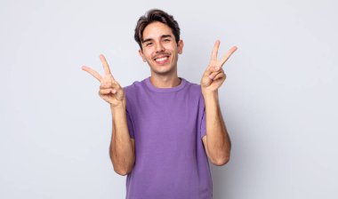 Genç, yakışıklı İspanyol adam gülümsüyor ve mutlu görünüyor, arkadaş canlısı ve memnun, iki eliyle zafer veya barış işareti yapıyor.