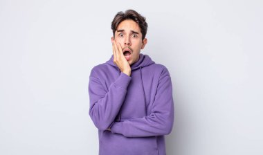 Genç, yakışıklı İspanyol bir adam şok ve inançsızlıkla ağzı açık, elleri yanağında ve kolları çaprazlanmış, sersemlemiş ve şaşırmış hissediyor.