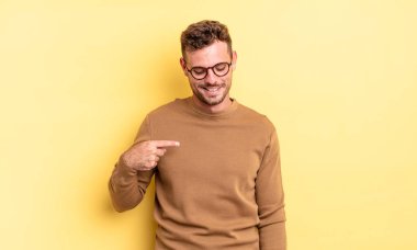 Genç, yakışıklı İspanyol adam neşeyle gülümsüyor, yere bakıyor ve göğsüne işaret ediyor.