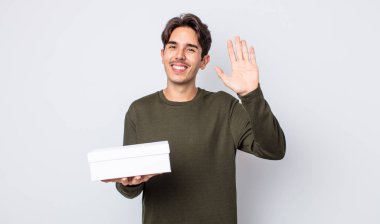 Genç İspanyol adam mutlu bir şekilde gülümsüyor, el sallıyor, sizi selamlıyor ve selamlıyor. beyaz kutu kavramı
