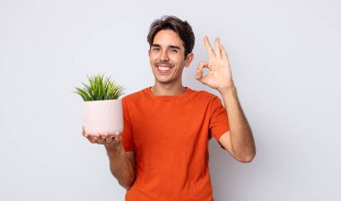 Genç İspanyol adam kendini mutlu hissediyor, iyi bir jestle onaylıyor. dekoratif bitki konsepti