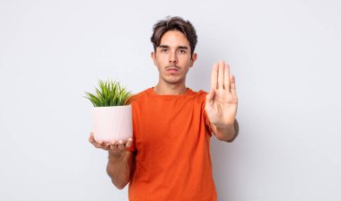 Genç İspanyol adam ciddi görünüyor. Avuç içi gösteriyor. Durdurma hareketi yapıyor. dekoratif bitki konsepti