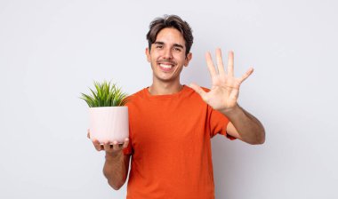 Genç İspanyol adam gülümsüyor ve arkadaş canlısı görünüyor, beş numarayı gösteriyor. dekoratif bitki konsepti