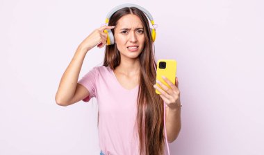 Genç ve güzel bir kadın kafası karışık ve şaşkın hissediyor, deli olduğunu gösteriyor. Kulaklık ve akıllı telefon.