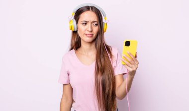 Genç, güzel bir kadın üzgün, üzgün ya da kızgın hissediyor ve yan tarafa bakıyor. Kulaklık ve akıllı telefon.