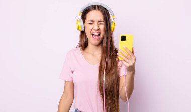 Genç ve güzel bir kadın agresif bir şekilde bağırıyor, çok kızgın görünüyor. Kulaklık ve akıllı telefon.