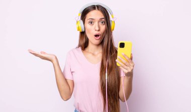 Genç ve güzel bir kadın şaşırmış ve şok olmuş görünüyor. Çenesi düşmüş ve elinde bir nesne tutuyor. Kulaklık ve akıllı telefon.