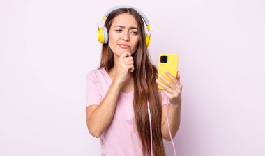 Genç, güzel bir kadın düşünüyor, kuşkulu ve şaşkın hissediyor. Kulaklık ve akıllı telefon.