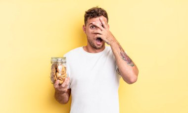 Genç yakışıklı adam şok olmuş, korkmuş ya da korkmuş görünüyor, yüzünü el kurabiyesi şişesi konseptiyle kapatıyor.