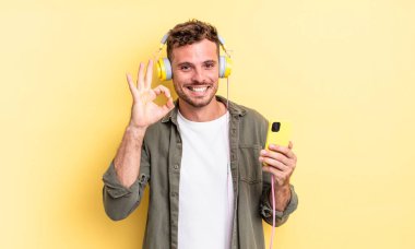 Genç yakışıklı adam kendini mutlu hissediyor. Onaylıyor. Jest kulaklıkları ve akıllı telefon konseptiyle.