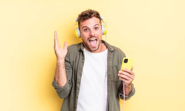 Genç yakışıklı adam kendini mutlu hissediyor ve inanılmaz bir kulaklık ve akıllı telefon konseptine hayret ediyor.