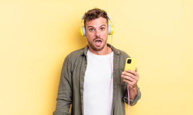 Genç yakışıklı adam şok olmuş ya da şaşırmış görünüyor. Kulaklıklar ve akıllı telefon konsepti.
